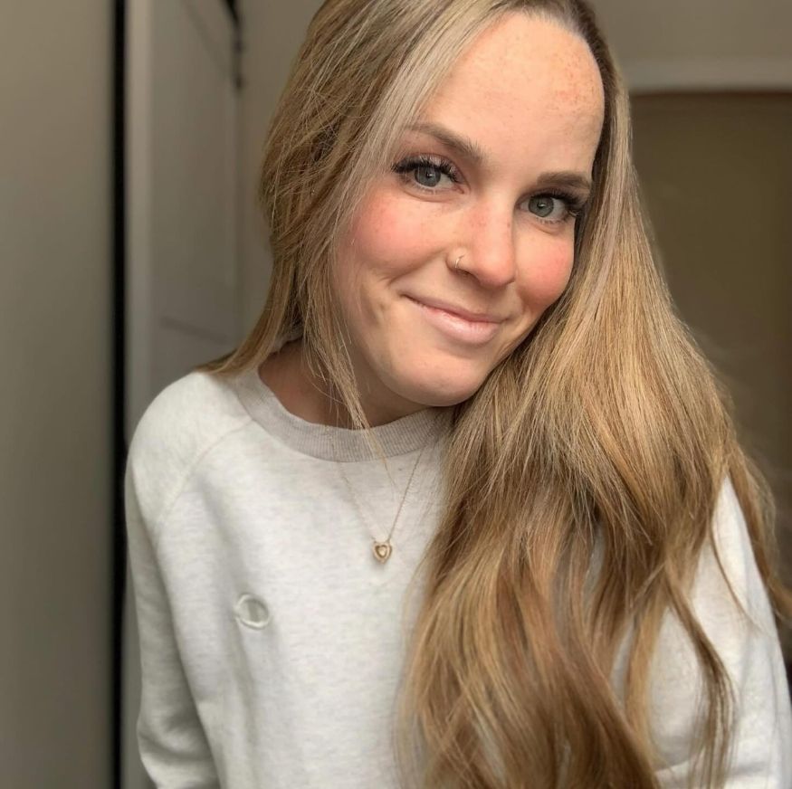 A smiling person with long blonde hair wearing a white sweater and delicate gold necklace in a casual indoor setting.
