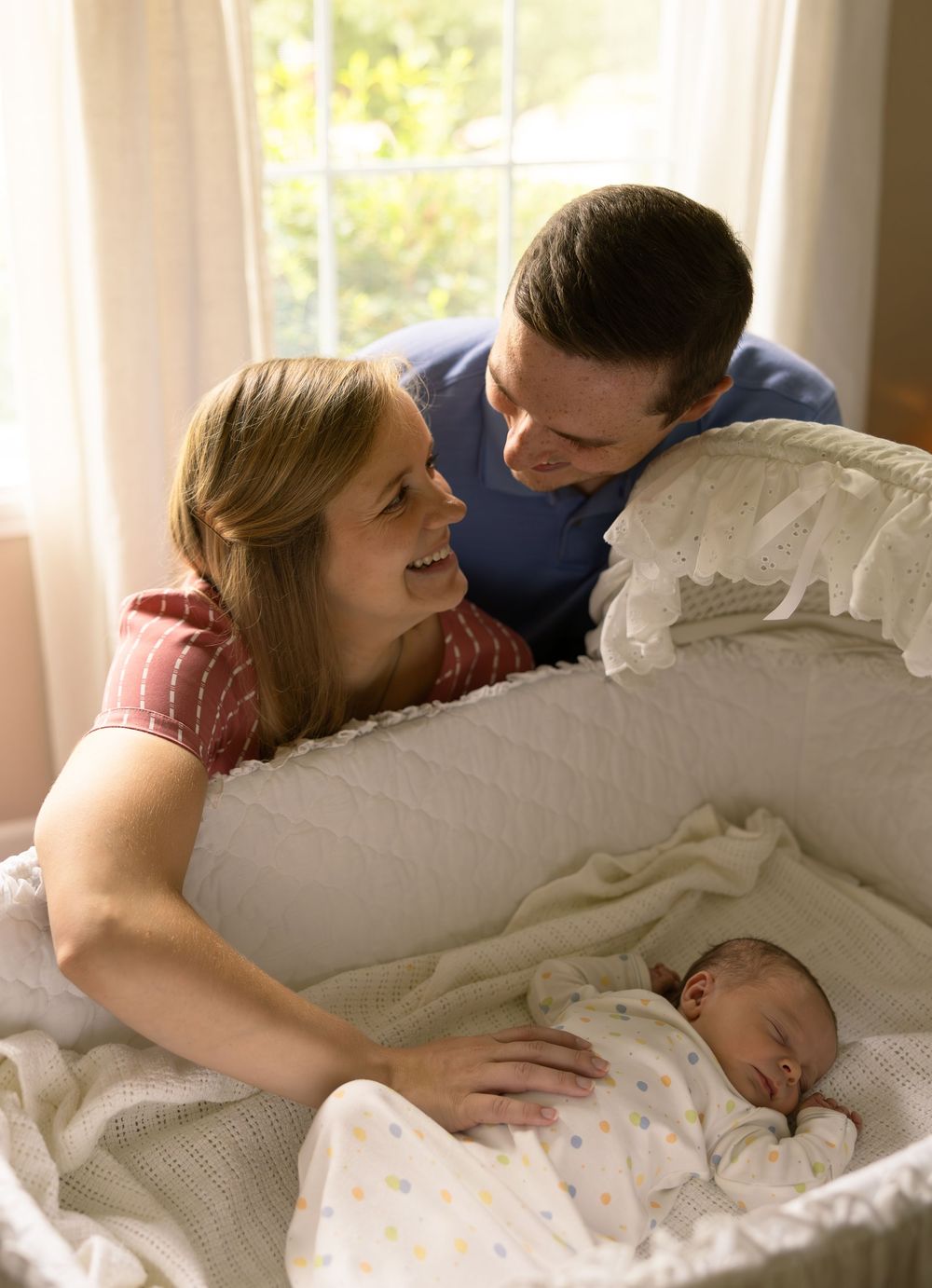 Parents lean over a white crib to look at their sleeping baby in a sunlit room.