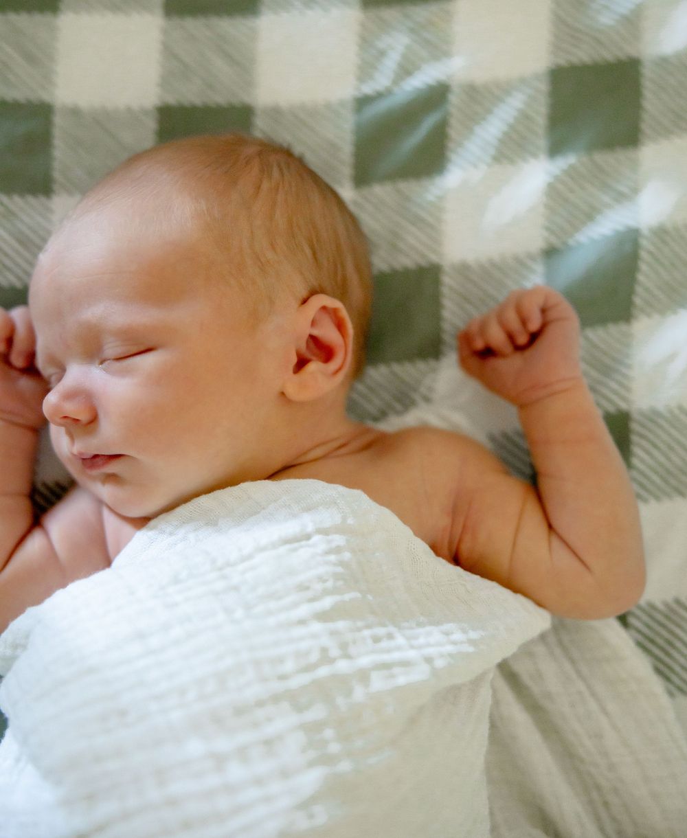 Sleeping newborn wrapped in white swaddle on buffalo check blanket.