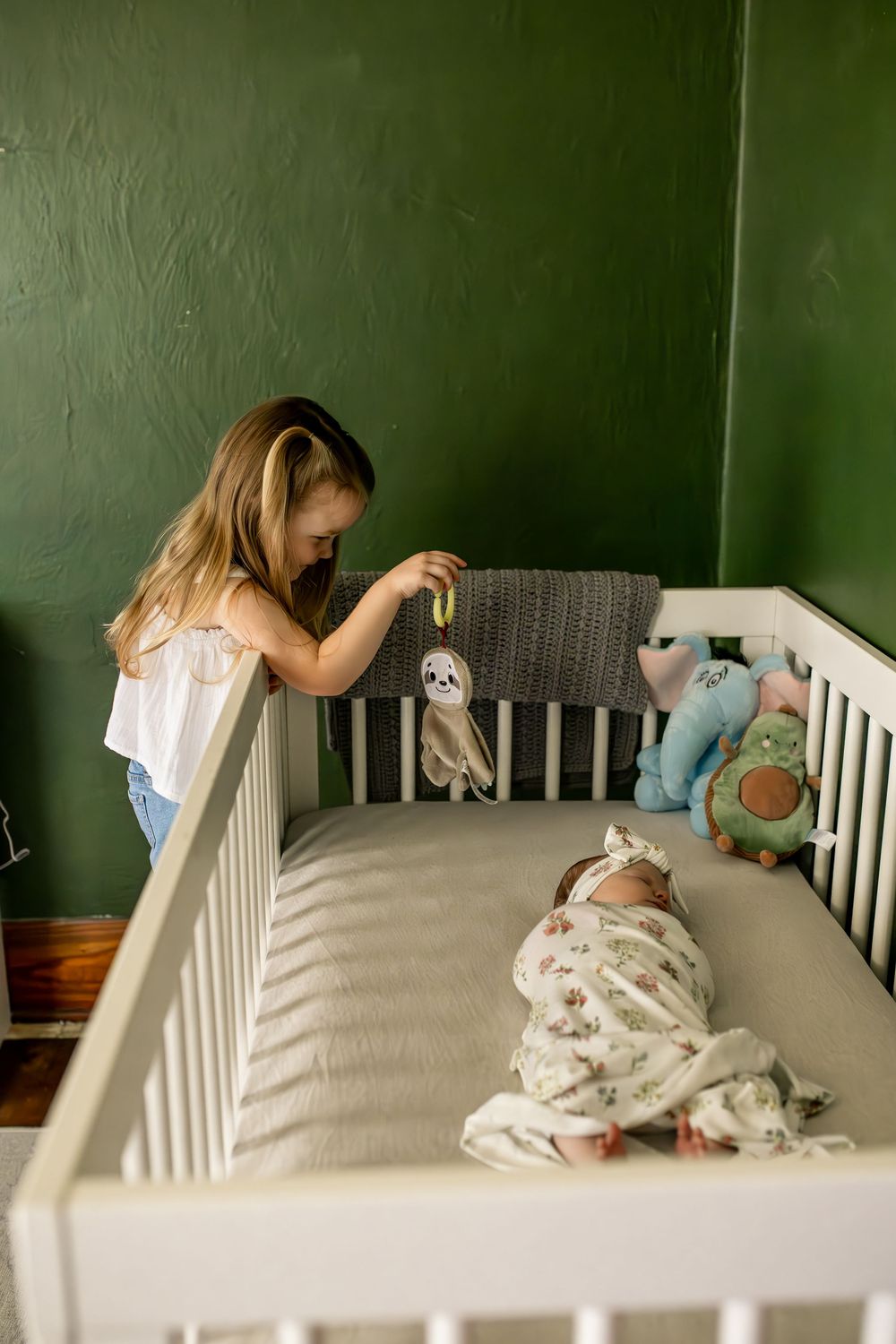A white crib with stuffed animals sits against a green wall in a nursery.