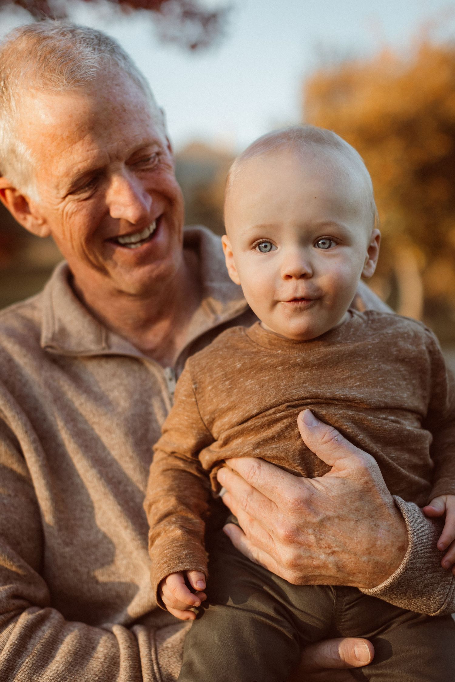 A smiling grandfather holds an infant boy in front of colorful fall foliage in golden-hour light.