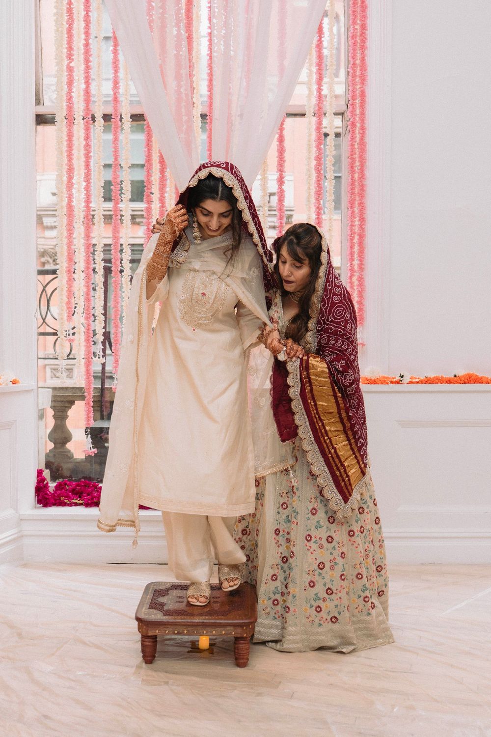 Two people in traditional Indian wedding attire performing a ceremonial ritual with pink curtains.