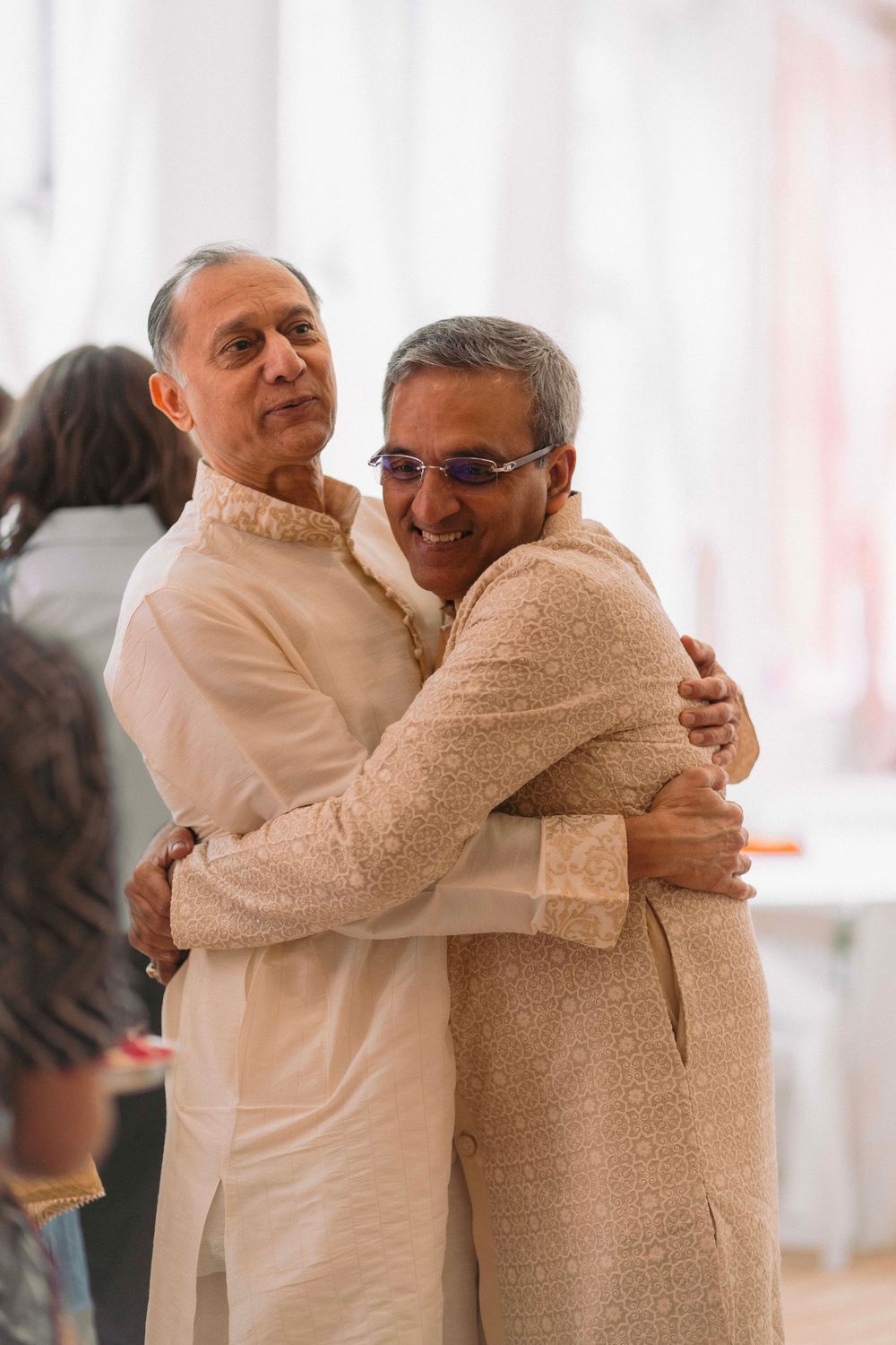 Two men in traditional cream-colored Indian attire sharing a warm embrace at a celebration.