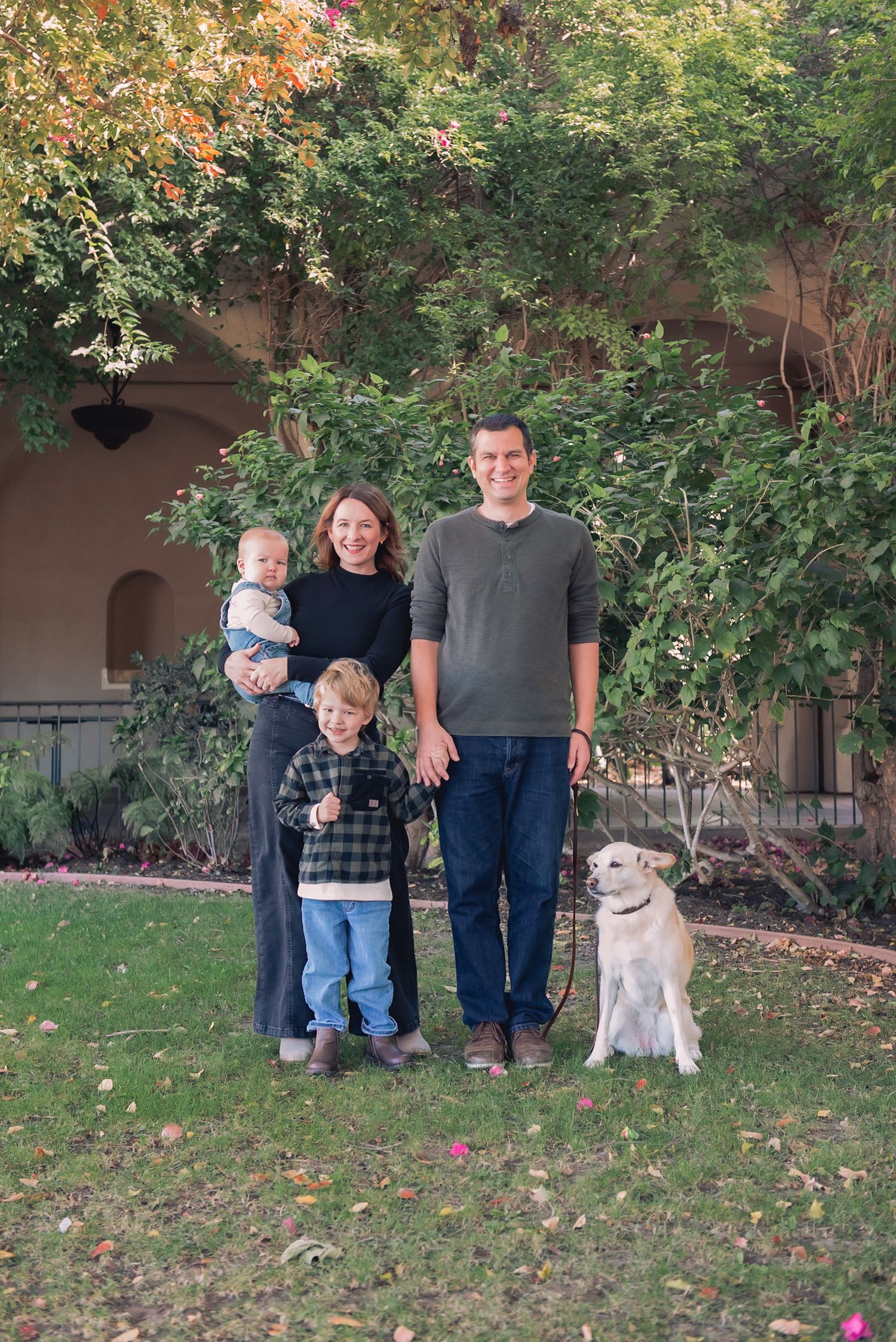 A group stands together with their friendly gray dog on a grassy lawn near autumn trees.