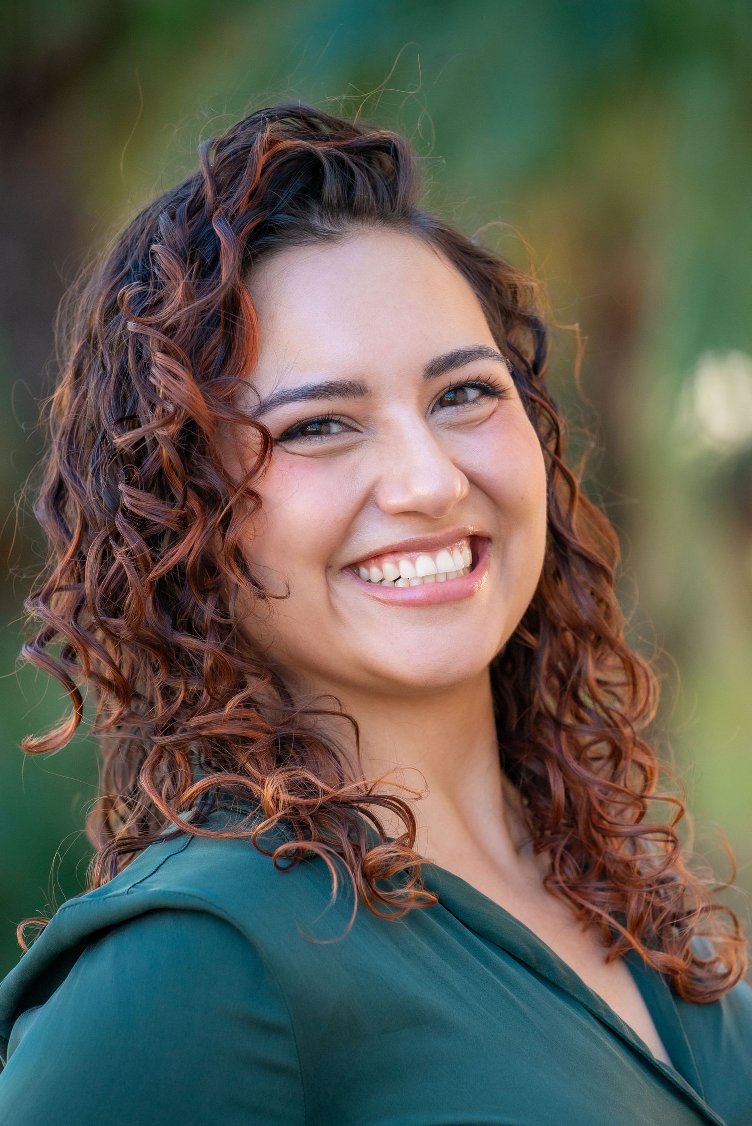 Portrait of someone with curly brown hair wearing a dark green top smiling warmly against a blurred outdoor background.