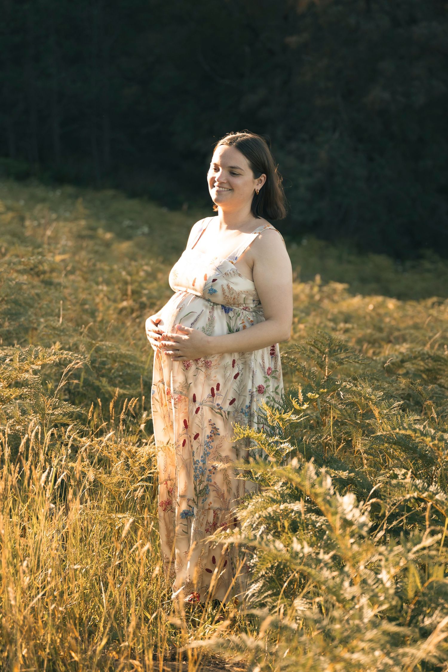 A pregnant woman in a floral dress stands in a sunlit meadow of golden grass during golden hour.