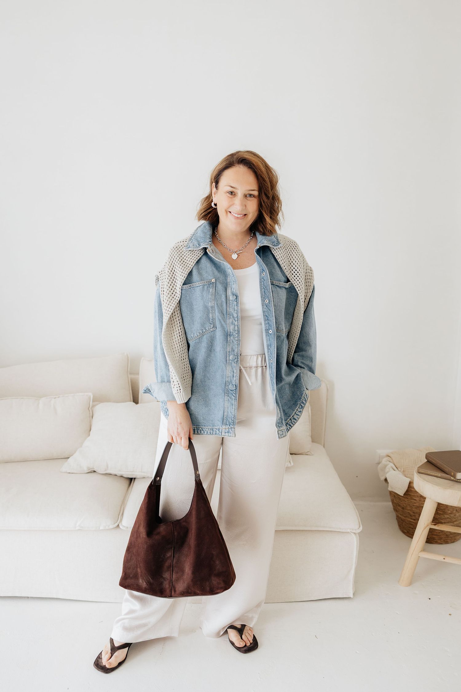 Model showcases casual outfit with light denim jacket, white pants and brown handbag in minimalist white setting.