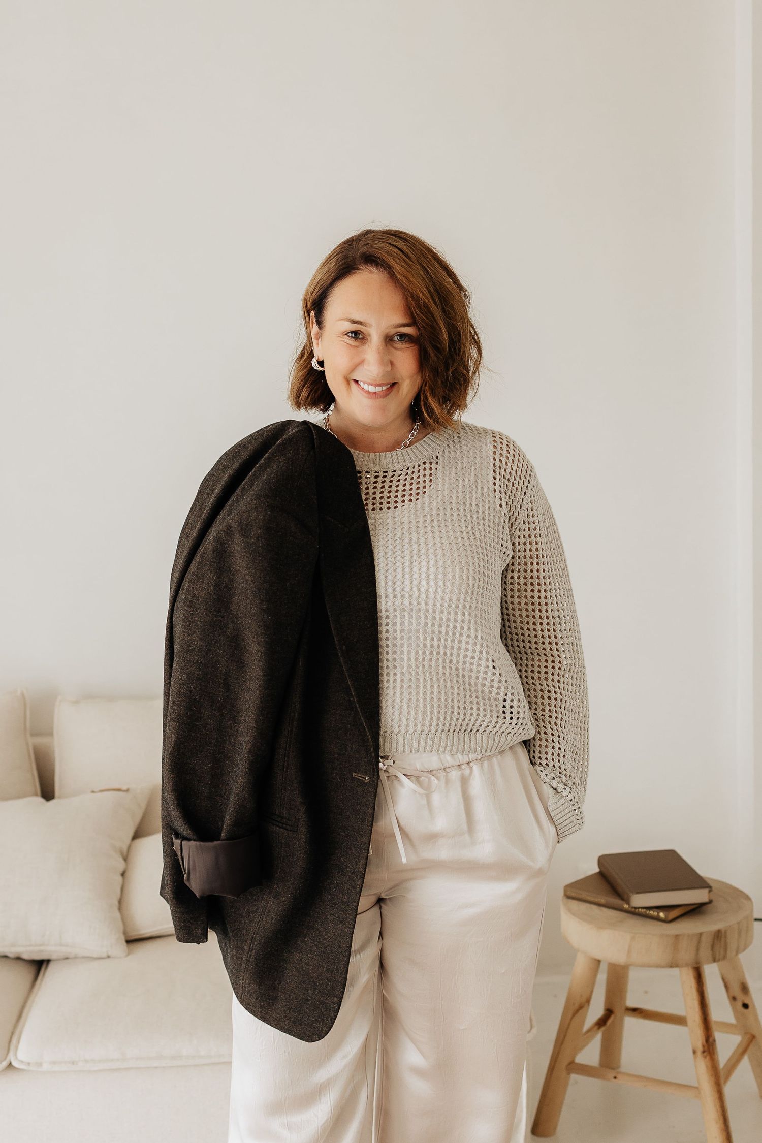 Casual lifestyle fashion shot showing a wavy-haired person in a knit sweater holding a black coat against white backdrop.