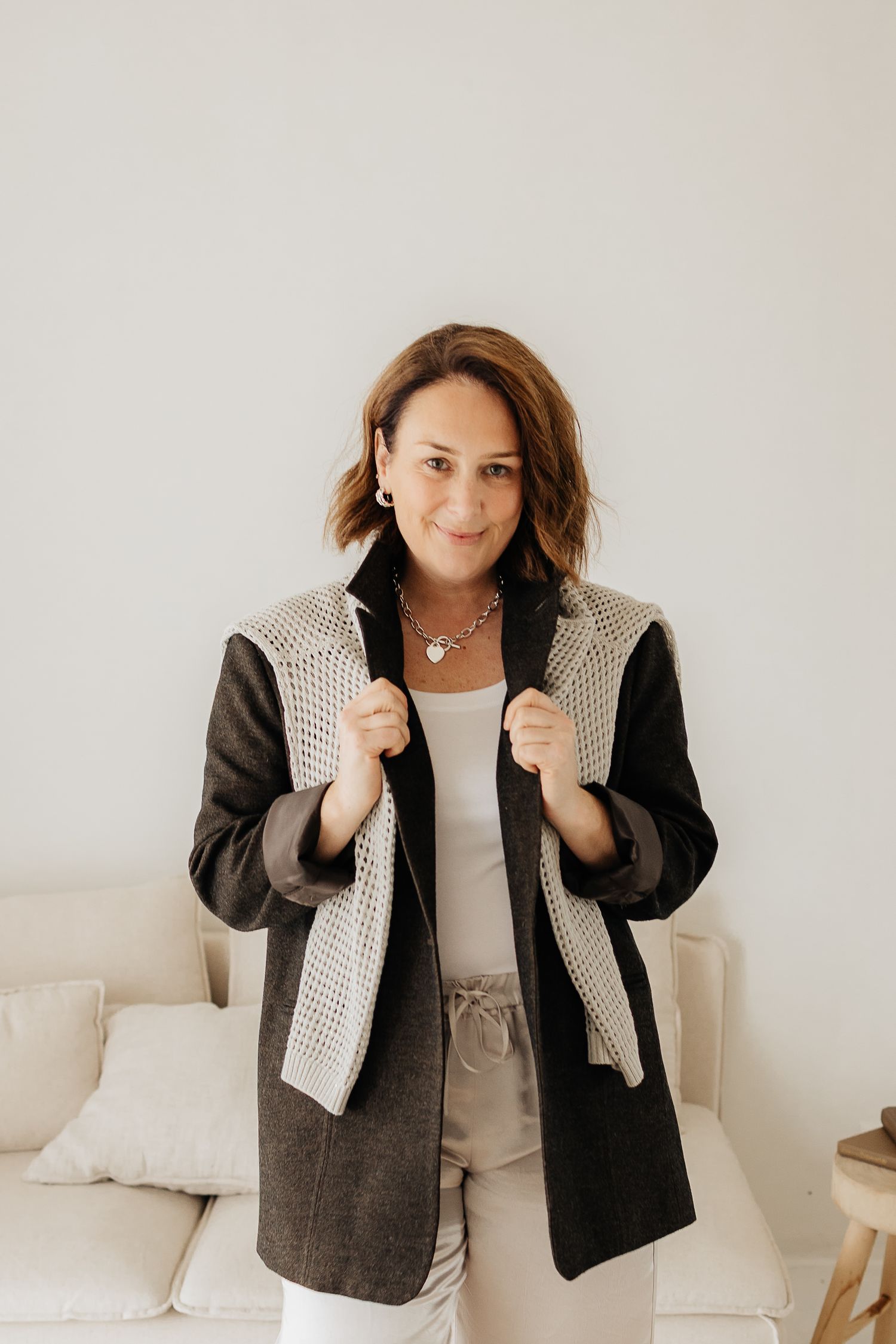 Model wearing black jacket with cream knit vest poses against white couch in lifestyle photoshoot.