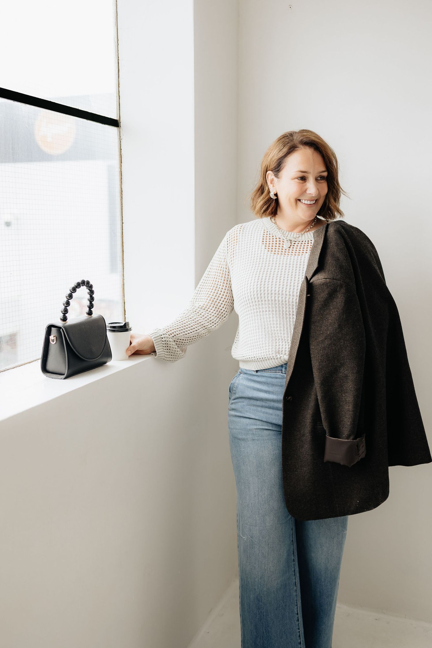 Woman in white sweater and jeans poses casually against white wall with black handbag.