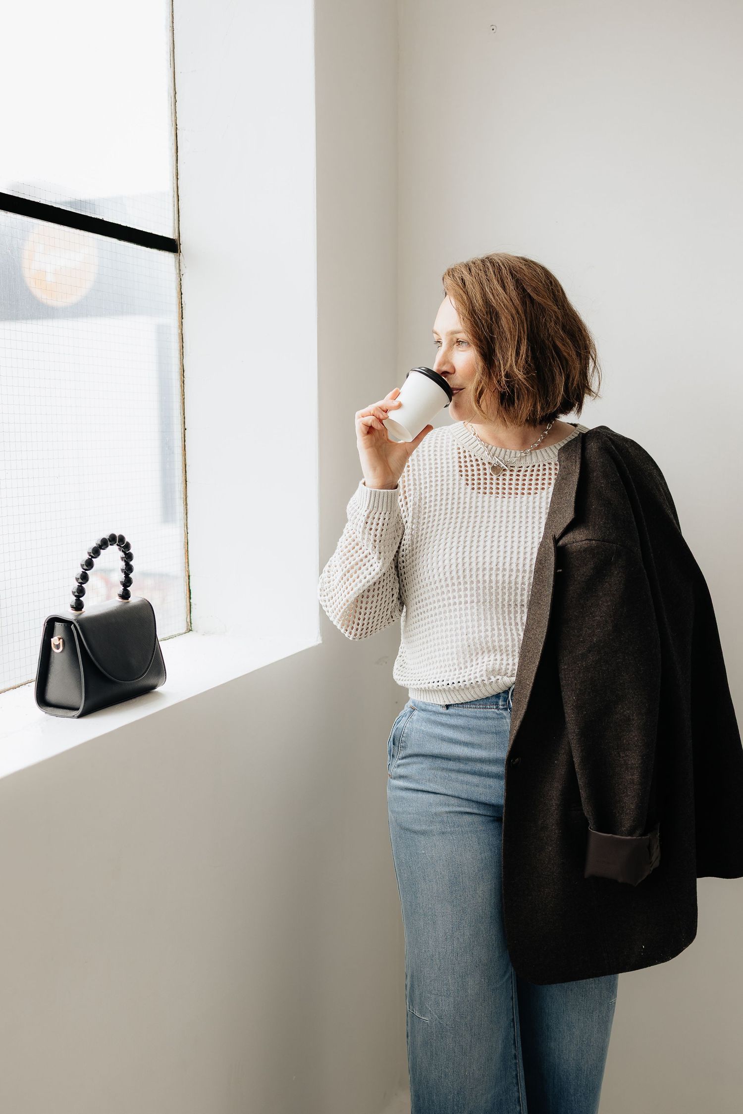 Lifestyle photo of someone in a cream sweater drinking coffee by a window with a black coat and handbag.