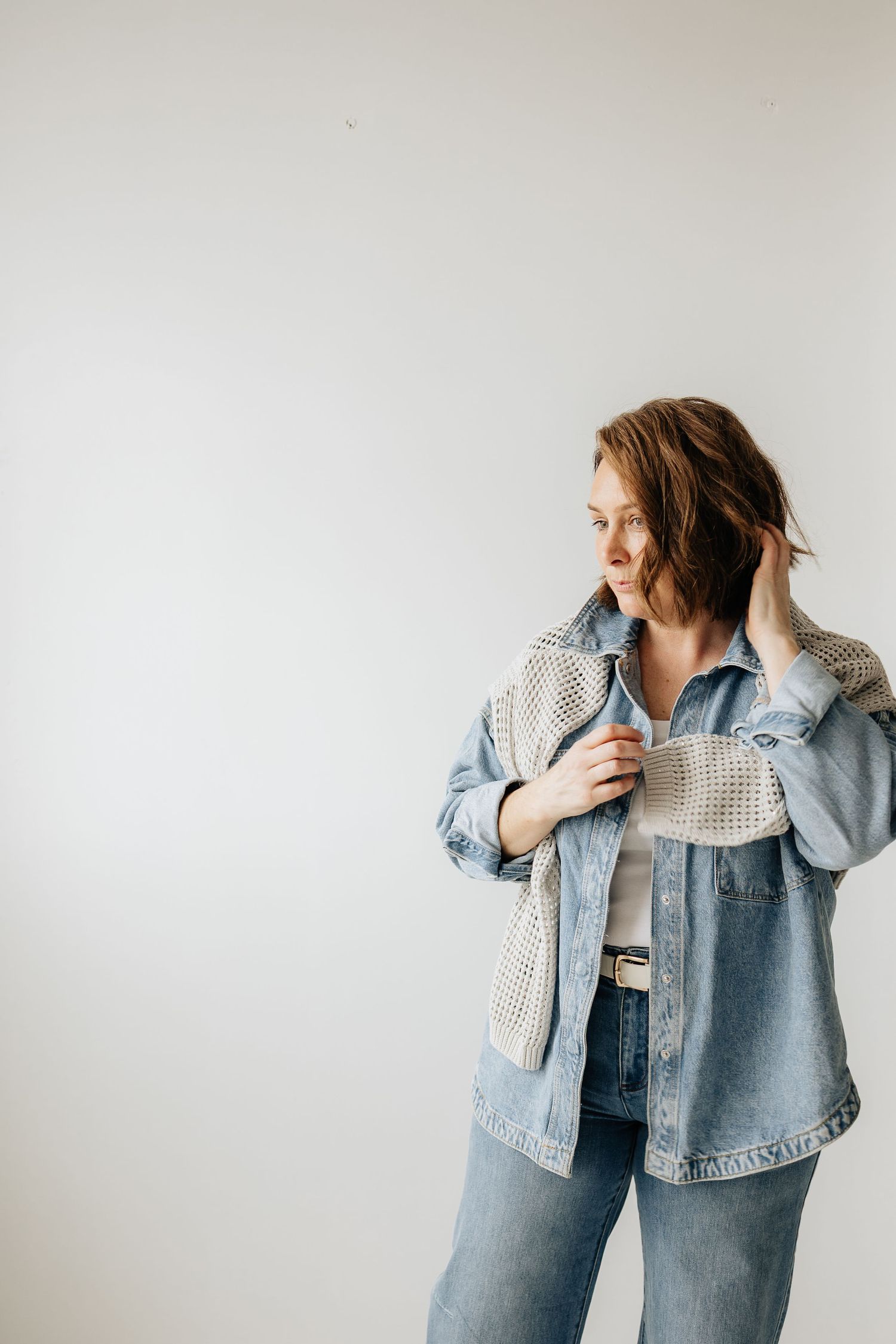 Model wearing light denim jacket and cream knit sweater poses against white wall showing casual style movement.