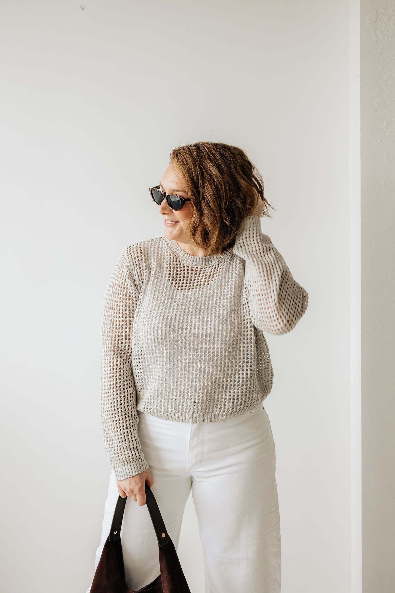 Person wearing a white mesh sweater and white skirt poses against a light background with a black handbag.