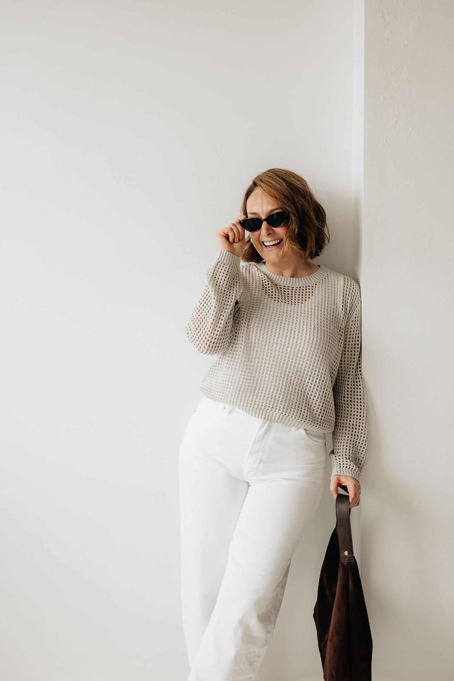 A person in a beige knit sweater and white pants poses against a white wall in a minimalist fashion series.