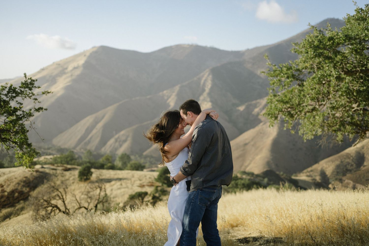 Couple embraces in golden mountain meadow during sunset with scenic mountain peaks in background.