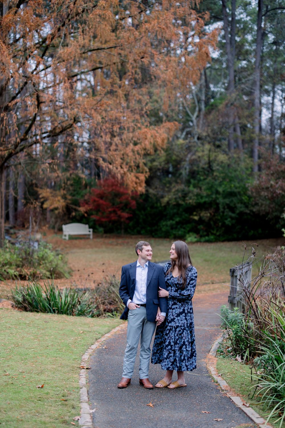 Couple walking along an autumn path with orange foliage and pine trees in the background.
