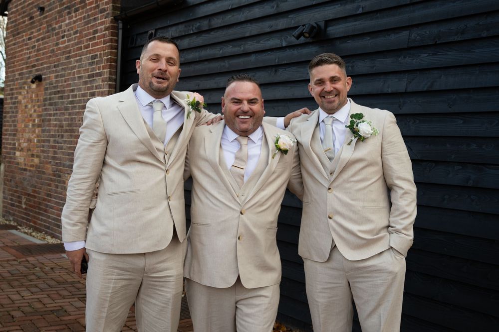 Groomsmen in beige suits pose together against a brick wall and black wooden siding backdrop.