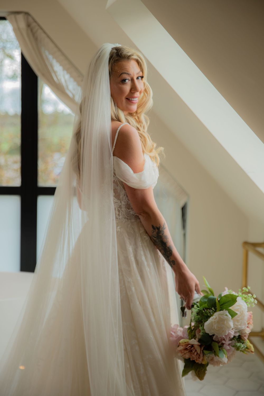 A bride in a white wedding gown and veil poses elegantly with her bouquet near a window.