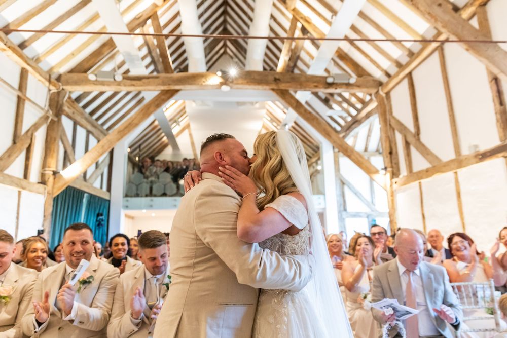 A romantic wedding ceremony moment captured in a sunlit barn with wooden beams and gathered guests.