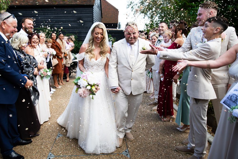 Wedding guests throw confetti as newlyweds walk down path outside venue in joyful celebration.