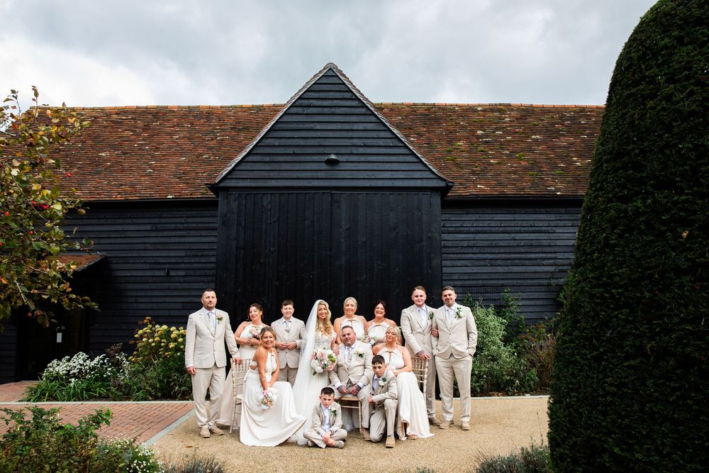 Wedding party dressed in white and beige poses together in front of rustic black barn buildings.