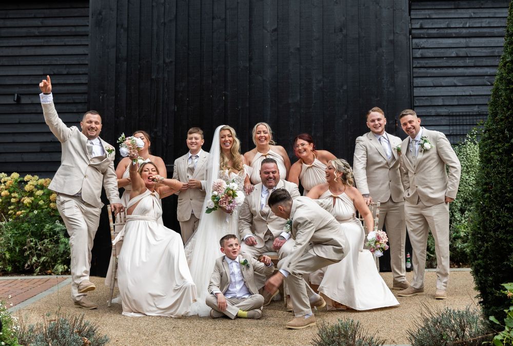 Wedding party in white and beige attire posing playfully against a dark barn wall.