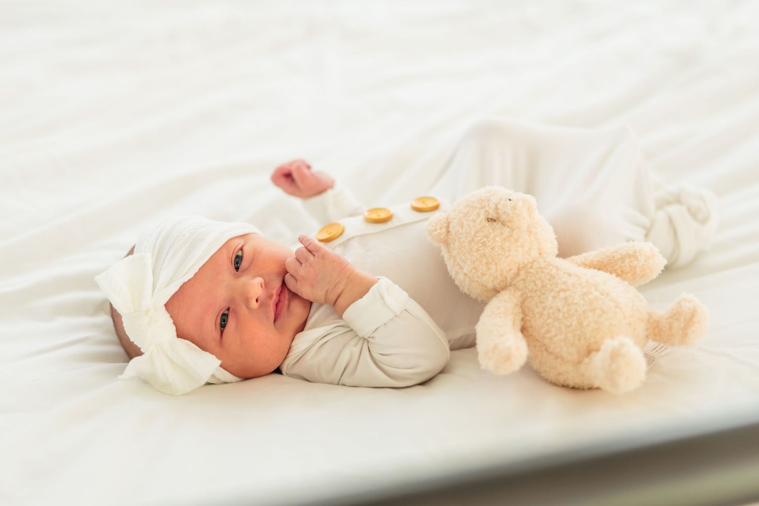 A baby in white clothing lies on a white blanket next to a plush teddy bear.