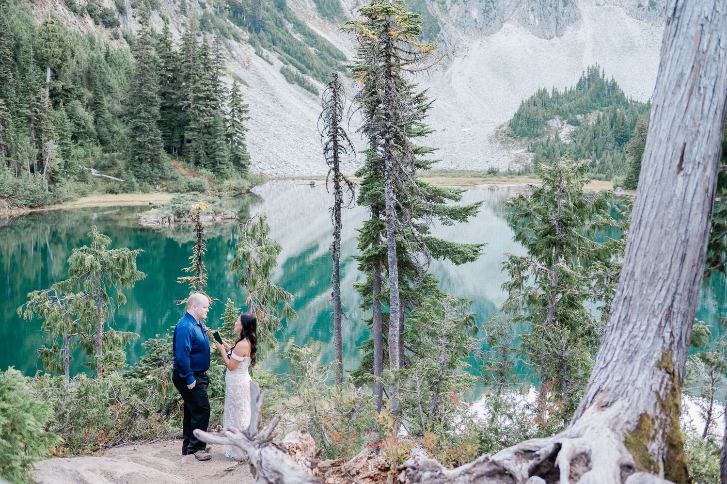 Couple exchanging vows in scenic mountain trail in Mt. Rainier National Park overlooking turquoise alpine lake surrounded by forest.