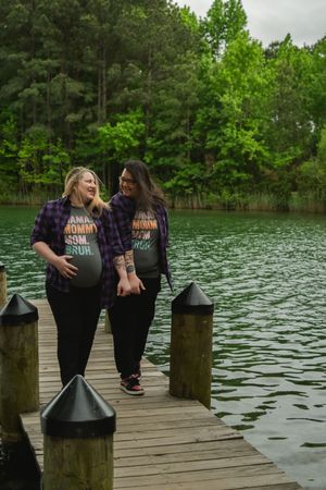 Two friends in matching outfits sit together on a wooden dock overlooking a serene lake surrounded by green trees.