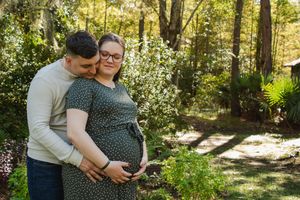 Maternity photo session in a sunlit forest with couple embracing among green trees and dappled sunlight.