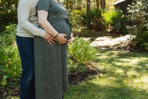 A loving embrace showing hands cradling a pregnant belly in a garden setting with sunlit greenery in the background.