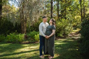 A couple walks together on a wooded trail surrounded by lush green trees and dappled sunlight on a sunny day.