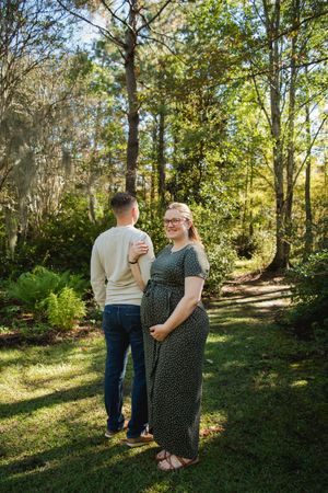 A couple stands together on a sunlit forest path surrounded by tall trees and lush greenery.