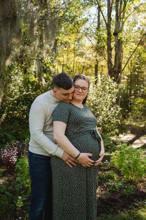 A couple embraces in a sunlit forest setting during a maternity photoshoot.