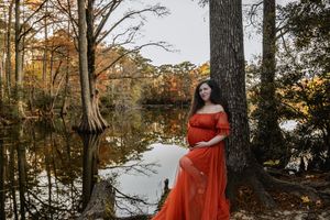 Figure in a flowing red dress stands among tall trees in a sunlit autumn forest setting.