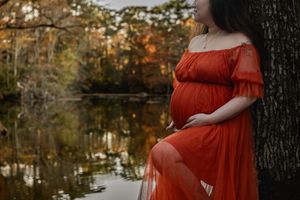 A vibrant red dress contrasts beautifully against dark tree trunks beside a reflective lake.