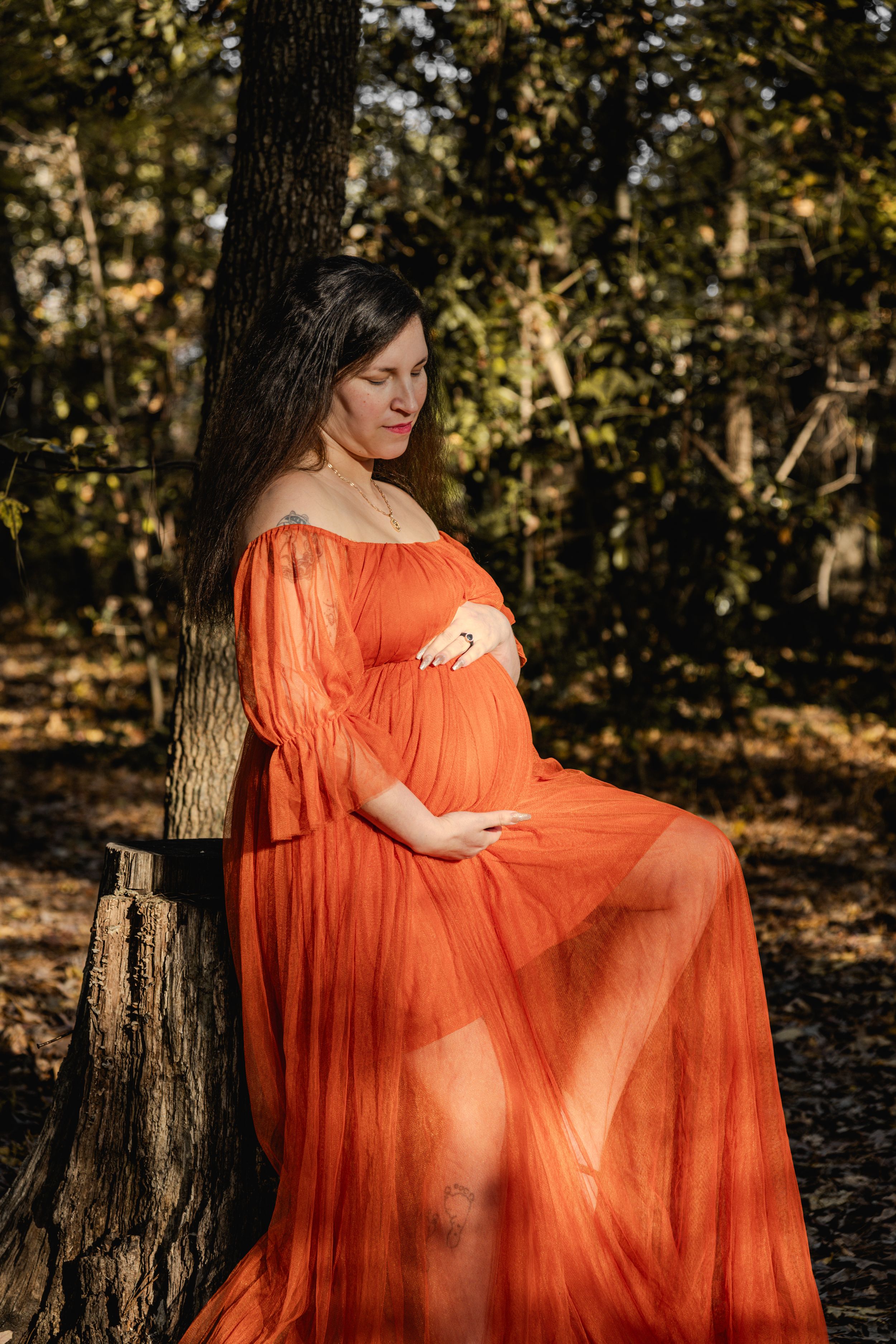 A flowing orange maternity dress catches sunlight in an autumn forest setting during a photoshoot.