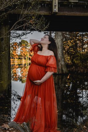 A figure in a flowing red dress stands by a reflective pond surrounded by autumn foliage.
