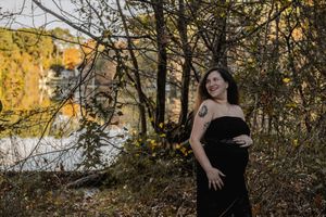 A person in a black dress poses outdoors during golden hour near a lake with autumn foliage in the background.