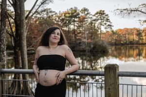 A person in a black dress poses thoughtfully by a serene lake with autumn trees in the background.