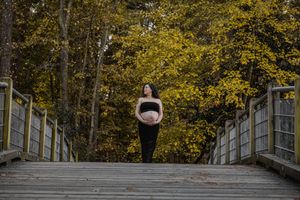 A pregnant woman stands on a wooden bridge surrounded by autumn trees with golden yellow foliage.