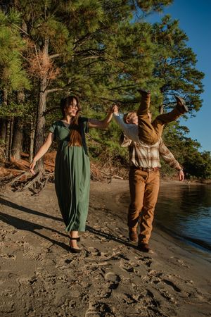Two people in vintage-inspired outfits dance joyfully along a scenic lakeside beach with pine trees in the background.