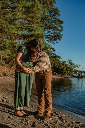 A couple shares an intimate moment along a rocky lakeshore with pine trees and blue water in the background.
