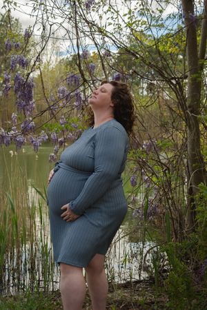 A person in a gray sweater dress poses outdoors near flowering wisteria and a lake in spring.