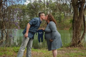 A couple shares a tender moment outdoors with a small blue outfit hanging between them near a wooded lake.