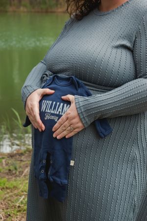 Pregnant woman in gray sweater dress holds tiny navy blue onesie with 'William' text by a lake.