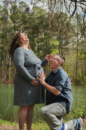 Couple shares playful moment by lake with trees in background during outdoor photo session.