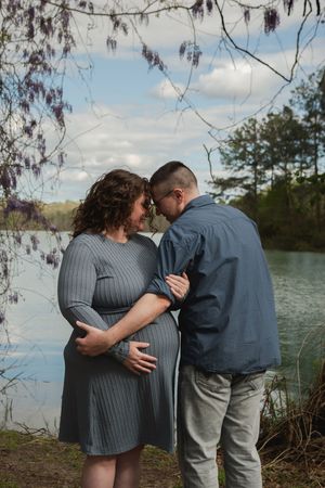 A couple embraces while standing by a serene lake on a sunny day with trees and clouds in the background.