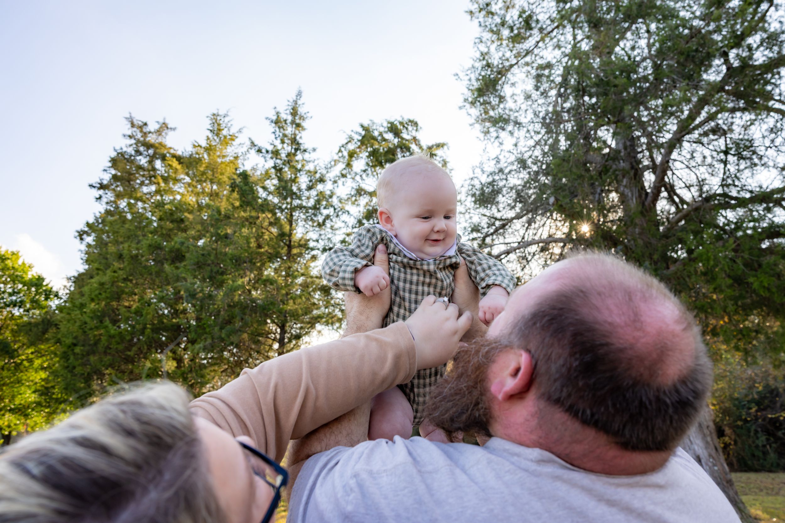A person lifts a small baby into the air while standing outdoors in a park setting during golden hour.
