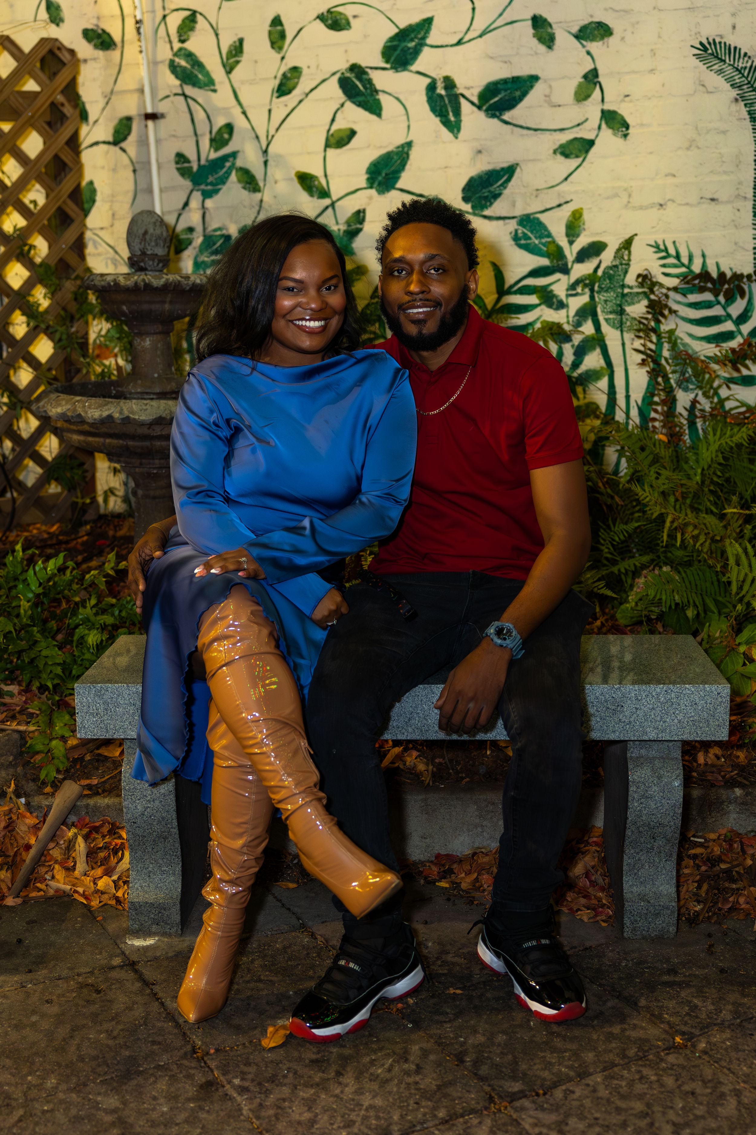 A couple sits together on a stone bench surrounded by lush greenery and foliage in an outdoor garden setting.